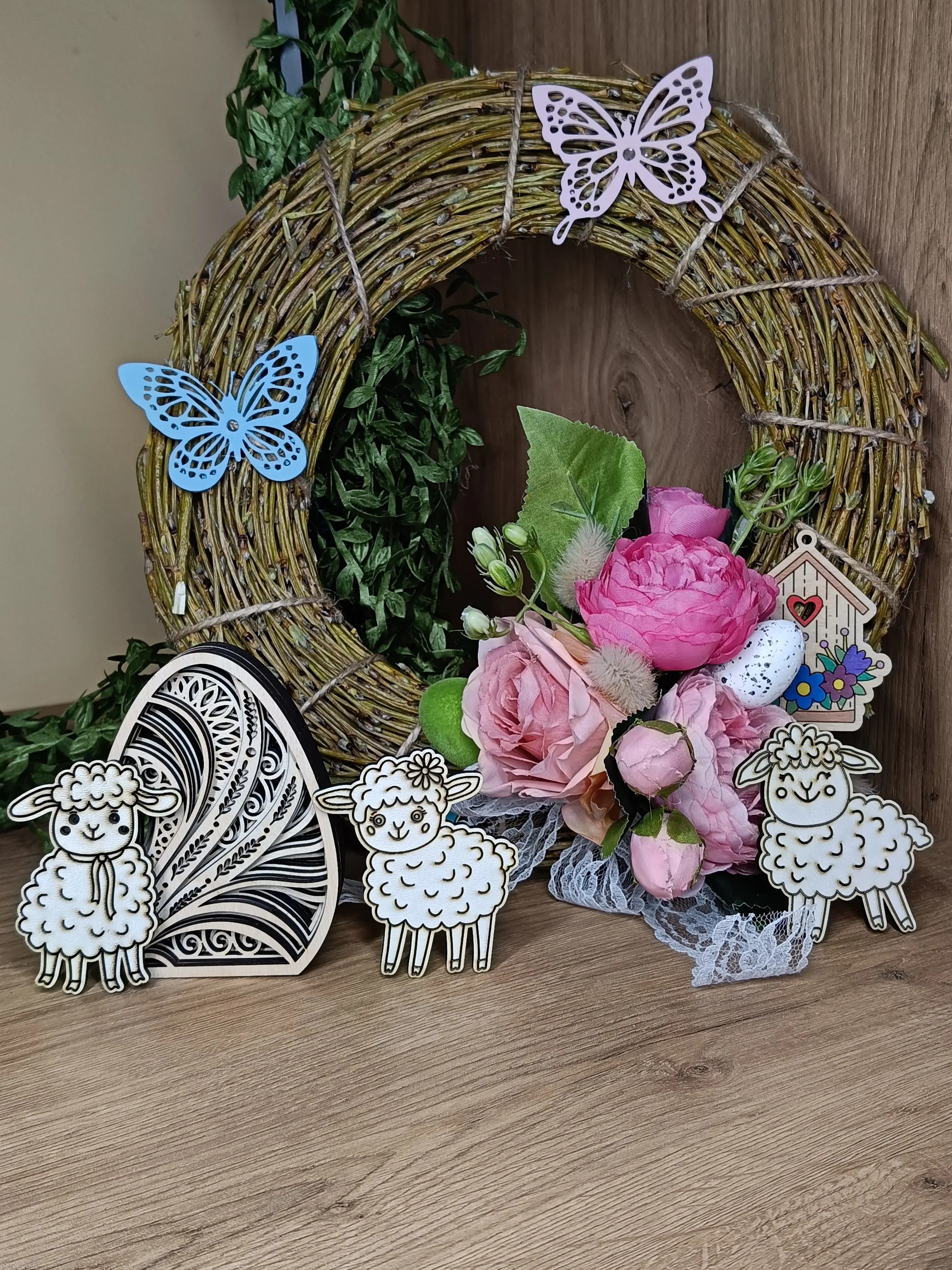 Rustykalna dekoracja wielkanocna z wiankiem, bukietem róż, trzema owieczkami oraz drewnianymi ozdobami w kształcie jajka i domku