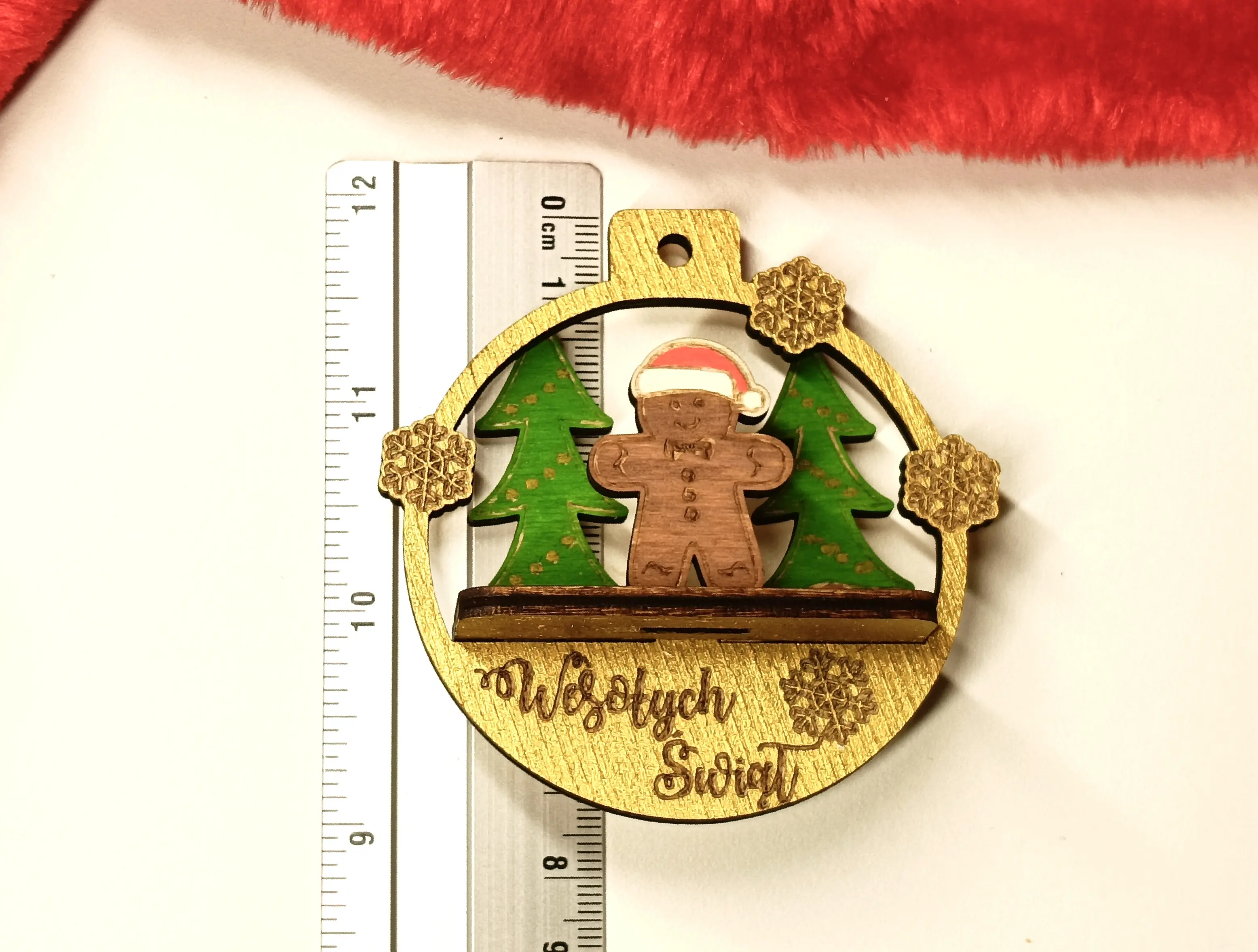 Drewniana bombka świąteczna z grawerem Wesołych Świąt, ozdobiona piernikowym ludzikiem w czapce Mikołaja, choinkami i złotymi śnieżynkami