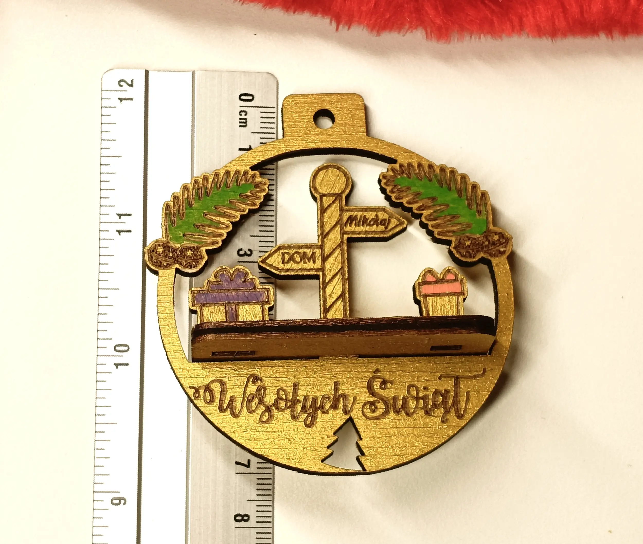 Drewniana bombka świąteczna z grawerem Wesołych Świąt, ozdobiona drogowskazem DOM i Mikołaj, prezentami oraz gałązkami sosny z szyszkami