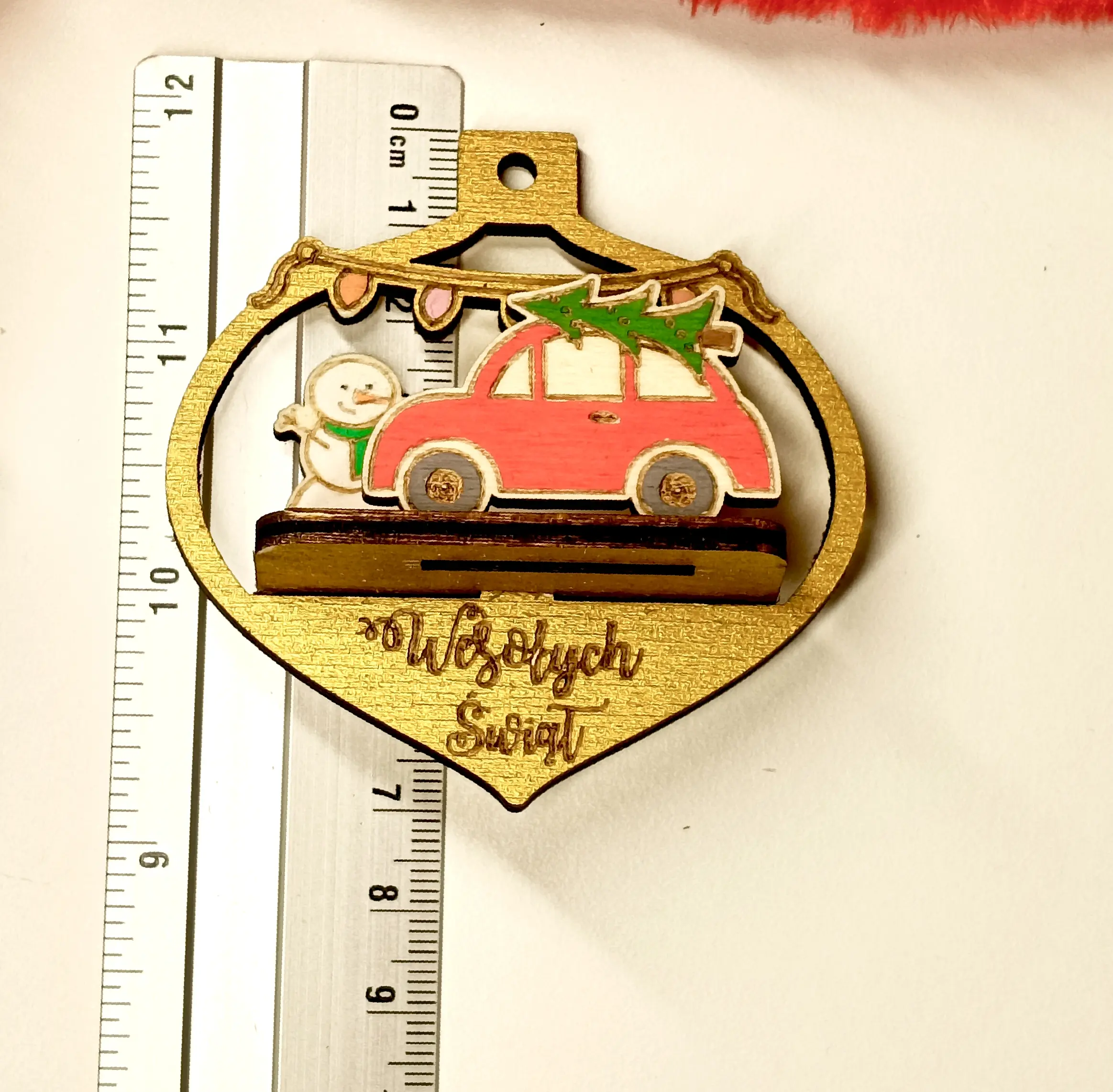Drewniana bombka świąteczna w kształcie łezki z grawerem Wesołych Świąt, ozdobiona czerwonym samochodem z choinką, bałwanem i kolorowymi lampkami