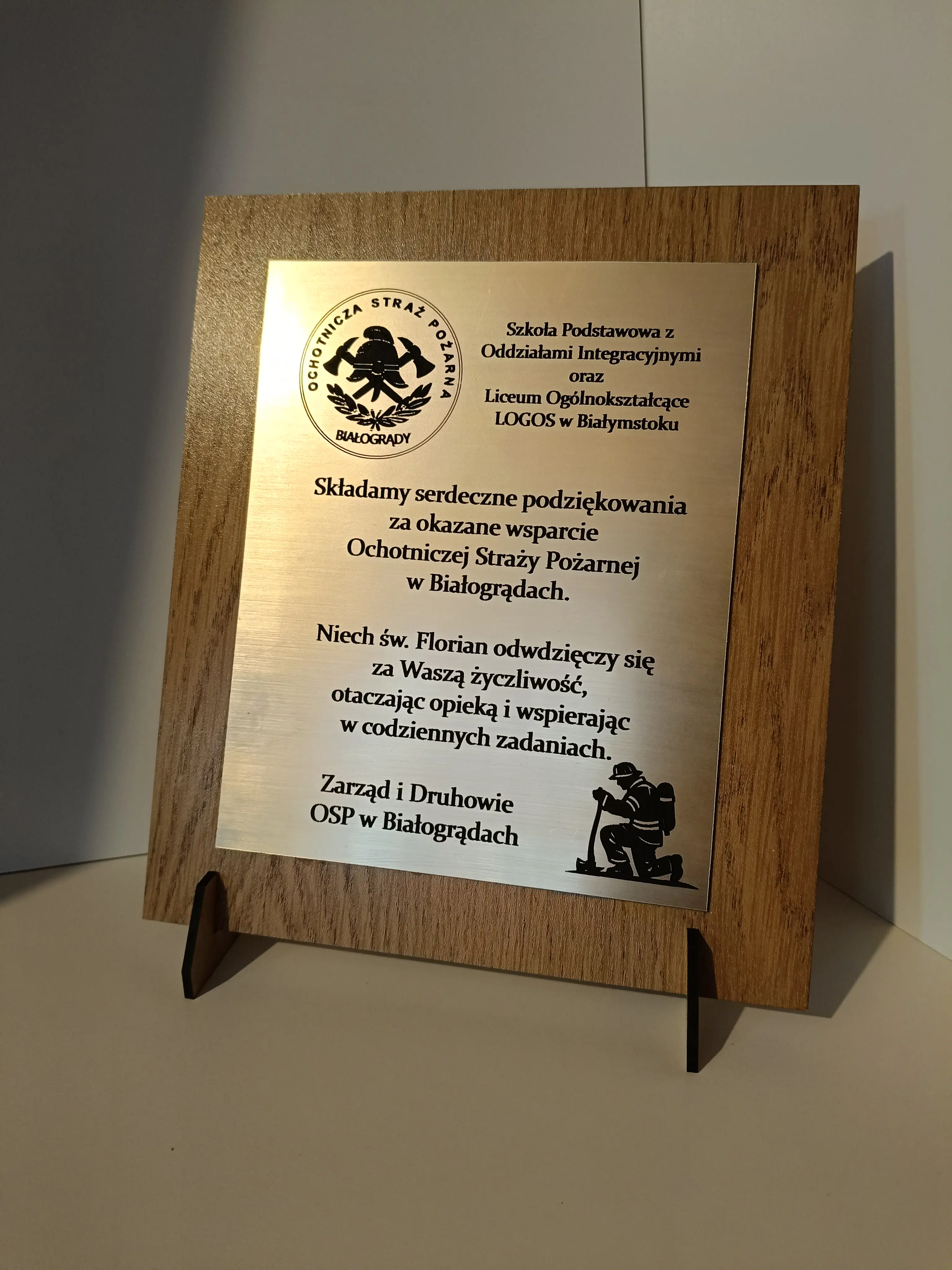 Drewniana tablica pamiątkowa na stojaku z grawerem i logotypem OSP Białogrądy, z dedykacją i grafiką strażacką