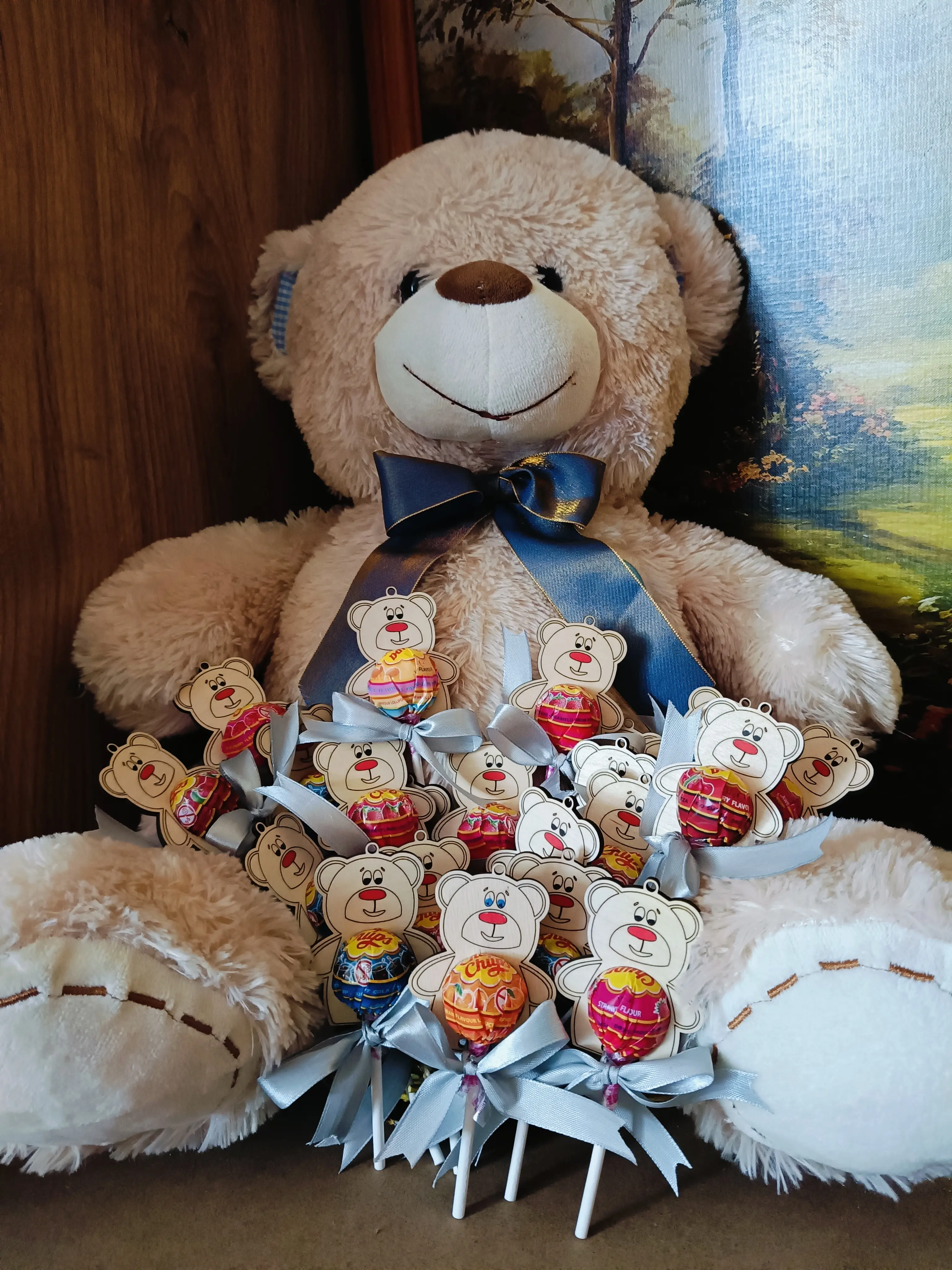 Pluszowy miś z niebieską wstążką trzymający bukiet lizaków ozdobionych buźkami misiów i kolorowymi wstążkami