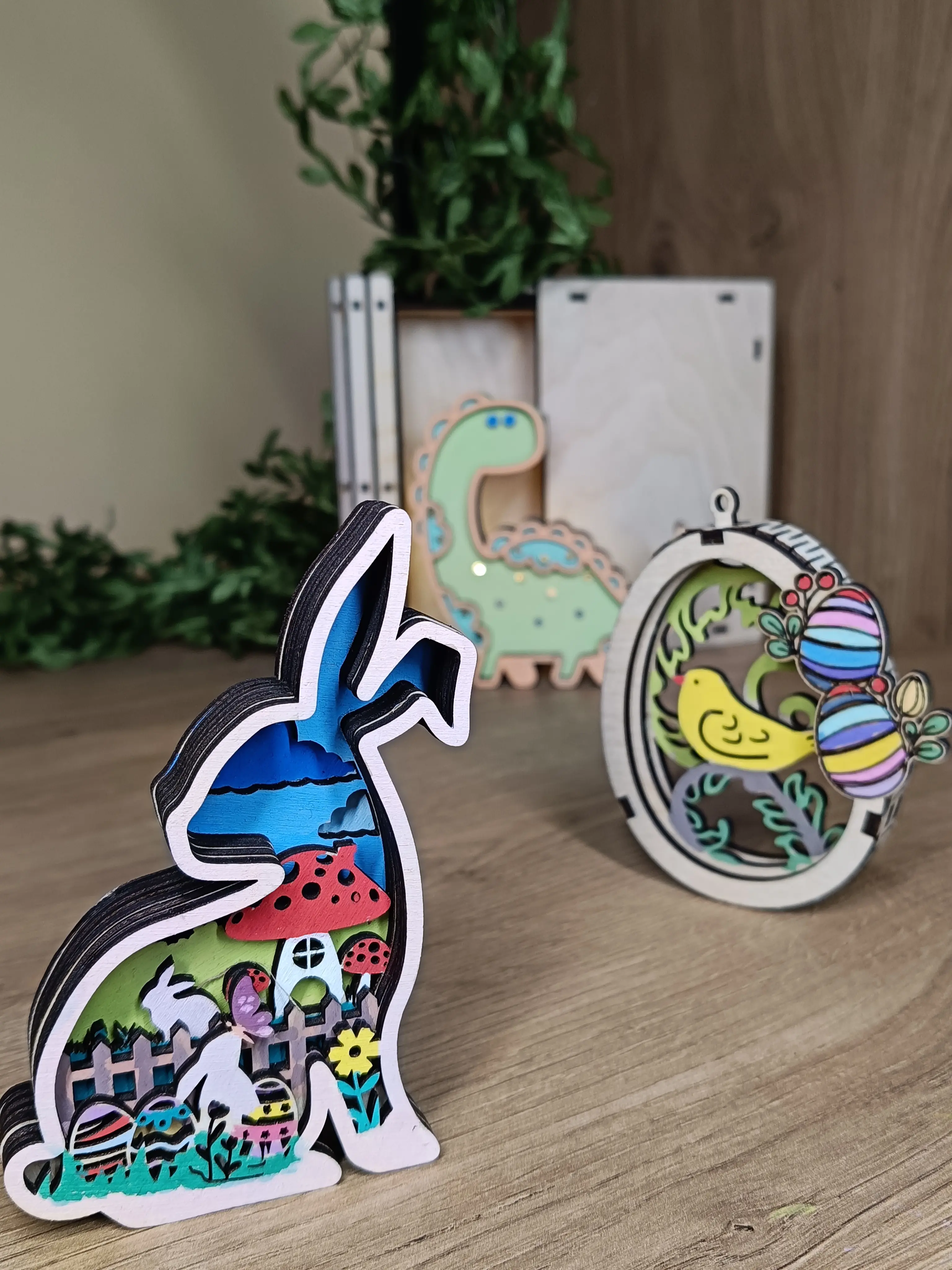 Kolorowe, warstwowe dekoracje drewniane przedstawiające królika ze scenerią, ozdobę z ptaszkiem i pisankami oraz pastelowego dinozaura